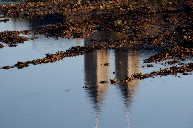 Weerspiegeling van de torens van de basiliek in Sint Odiliënberg. De torens spiegelen in een plas water. In de plas liggen ook oranje herfstbladeren.