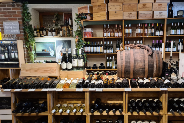 Een winkel met schappen vol flessen wijn. Bovenop de schappen staan kratten. In het midden is een wijnvat zichtbaar.