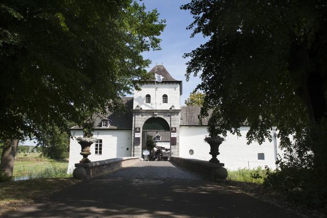 We zien een wit gebouw met een torentje en een grote, ronde opening in de muur onder de toren. Er leidt een brug over het water naar de opening. Links en rechts staan bomen. Het is de entree van kasteel Daelenbroeck.