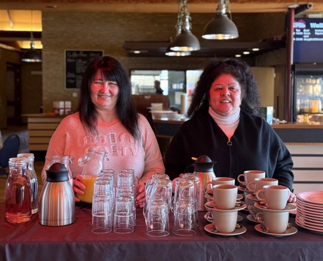 2 dames die in een restaurant-omgeving achter een tafel staan met daarop glazen en mokken