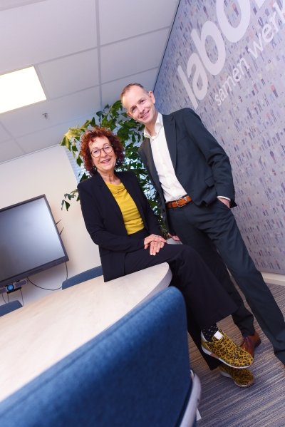 Twee personen in nette kleding staan in een kantooromgeving. Eén persoon zit op de rand van een tafel, de ander staat ernaast. Op de achtergrond zijn een grote kamerplant en een wand met een patroon en tekst zichtbaar. Het licht komt van plafondlampen in het kantoor.