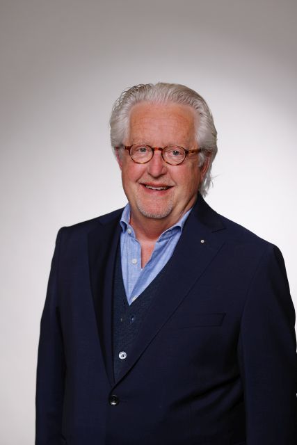 Persoon in een donker colbert en overhemd poseert voor een neutrale achtergrond