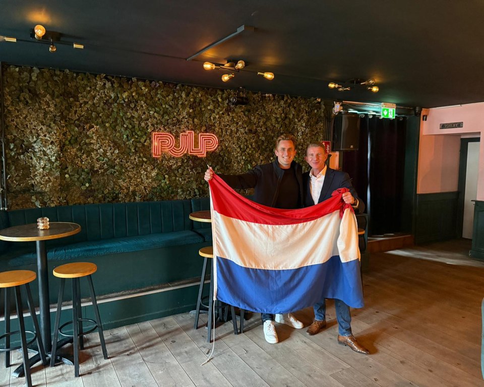 Burgemeester Jack de Vries (rechts) en Wiebe Kuiper, voorzitter van Koninklijk Horeca Nederland Maassluis (links), poseren samen met de Nederlandse vlag en onderstrepen de samenwerking rondom het WK Voetbal 2026.