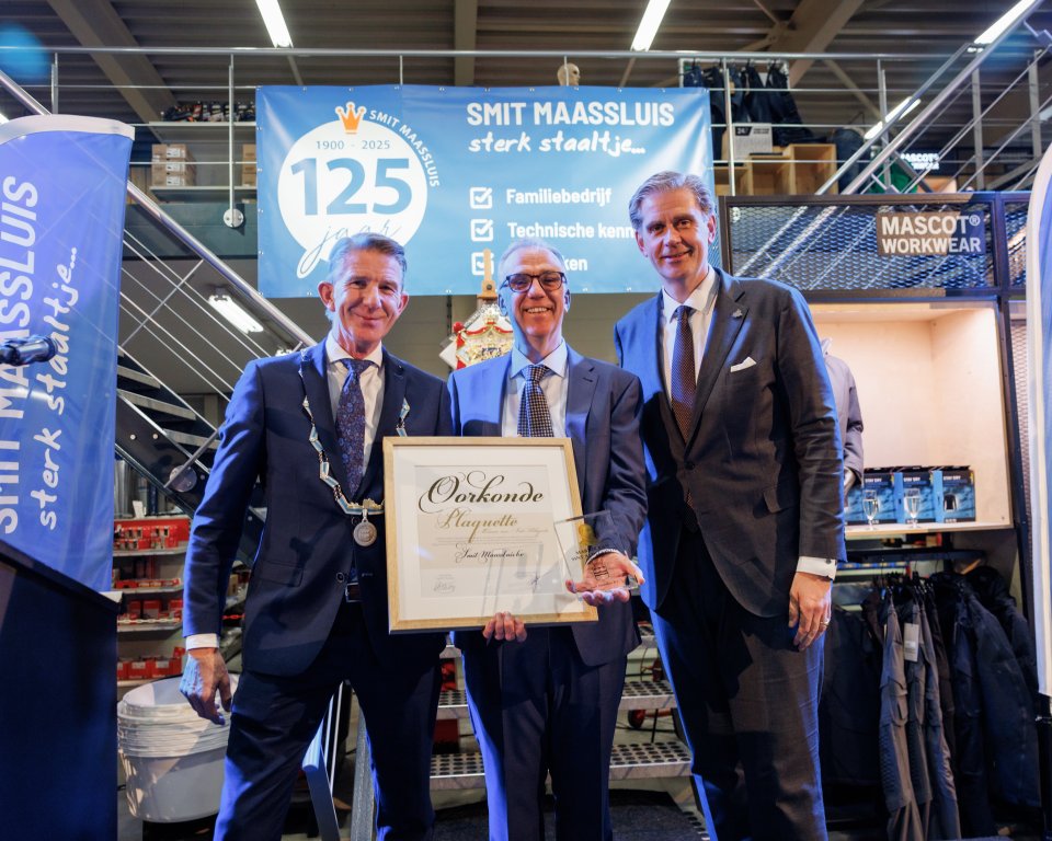 De foto toont dat burgemeester Jack de Vries de Marnix van Sint Aldegonde‑plaquette uitreikt aan Peter Smit, in het bijzijn van de Commissaris van de Koning in Zuid-Holland, als erkenning voor 125 jaar ondernemerschap en de bijzondere betekenis van Smit Maassluis voor de stad Maassluis.