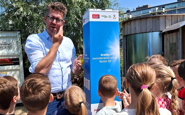 Foto van wethouder Kroonen die zich bij de zonnebrandpaal insmeert met zonnebrandcrème, leerlingen van IKC Het Balkon kijken toe en