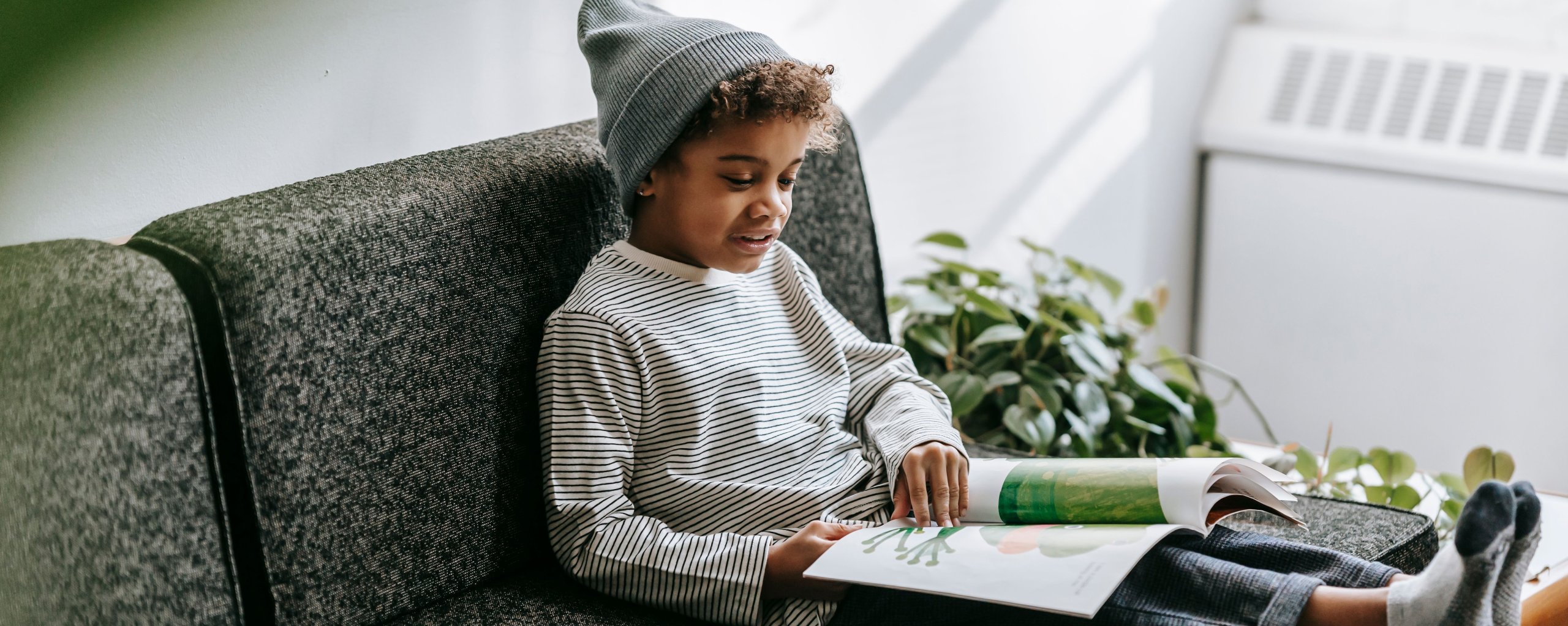 Een jonge jongen in een beanie en gestreept shirt leest een boek op een bank, omringd door planten.