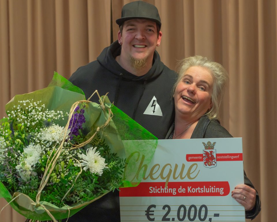 Steve Bergsma en Jolanda Ruitenbeek van Stichting de Kortsluiting houden een cheque van 2000 euro en een bos bloemen omhoog