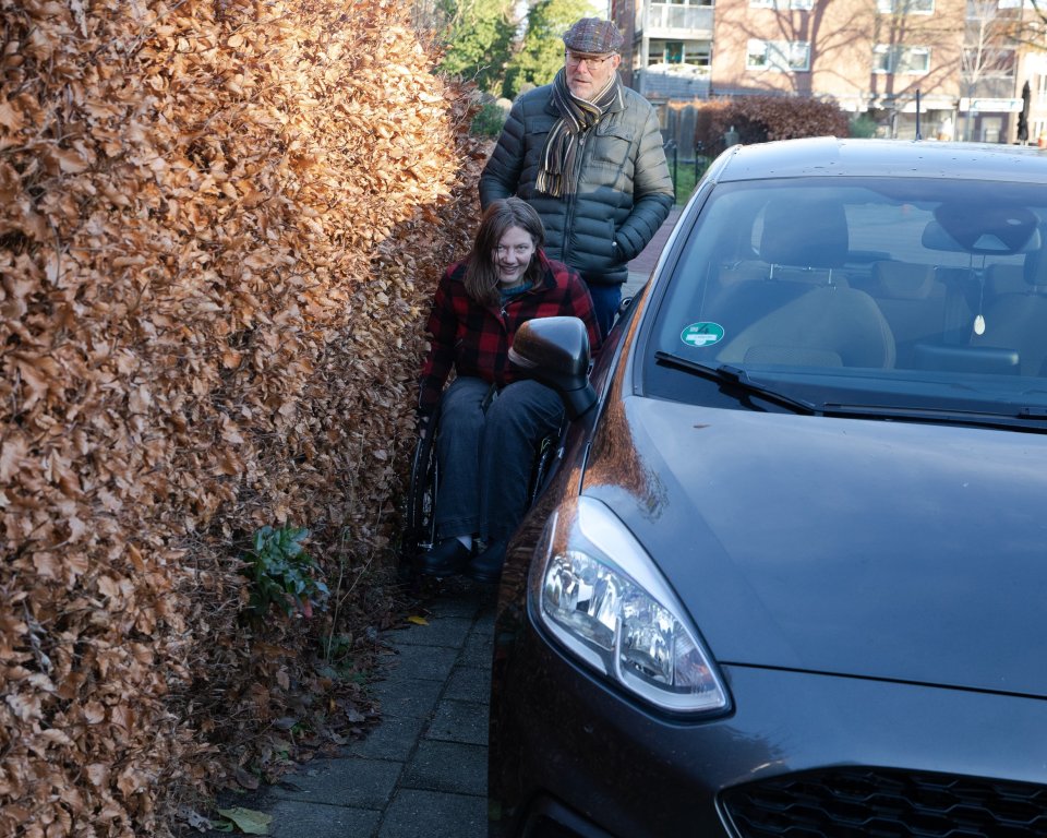 Persoon in rolstoel die niet langs een auto kan die op de stoep geparkeerd staat. 