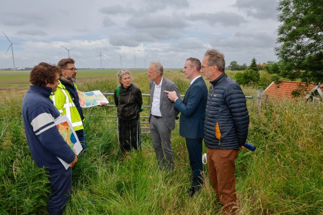 Je kijkt naar het gezelschap op de dijk, tussen het hoge groene gras. Op de achtergrond staan windmolens in een lager gelegen gebied. Op de foto staan v.l.n.r. historicus Lars Boon, archeoloog Sander Gerritsen, staf deltacommissaris Lilianne van Sprundel, Deltacommissaris Co Verdaas, burgemeester Herman Wiersema, Wethouder Herman ter Veen.