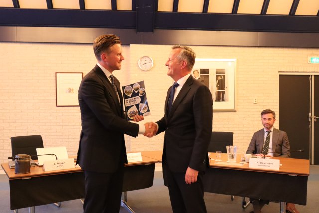 Foto waarop raadslid Bram Beemster de profielschets van de burgemeester overhandigt aan commissaris van de Koning Arthur van Dijk