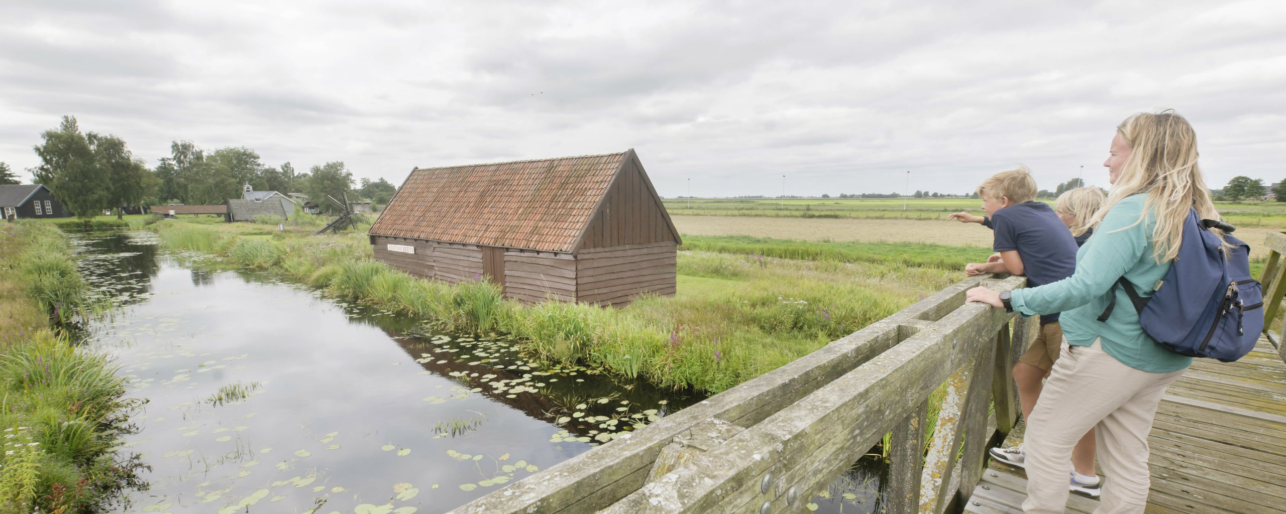 Moeder met kinderen kijken vanaf een brug uit over een sloot naar een schuurtje