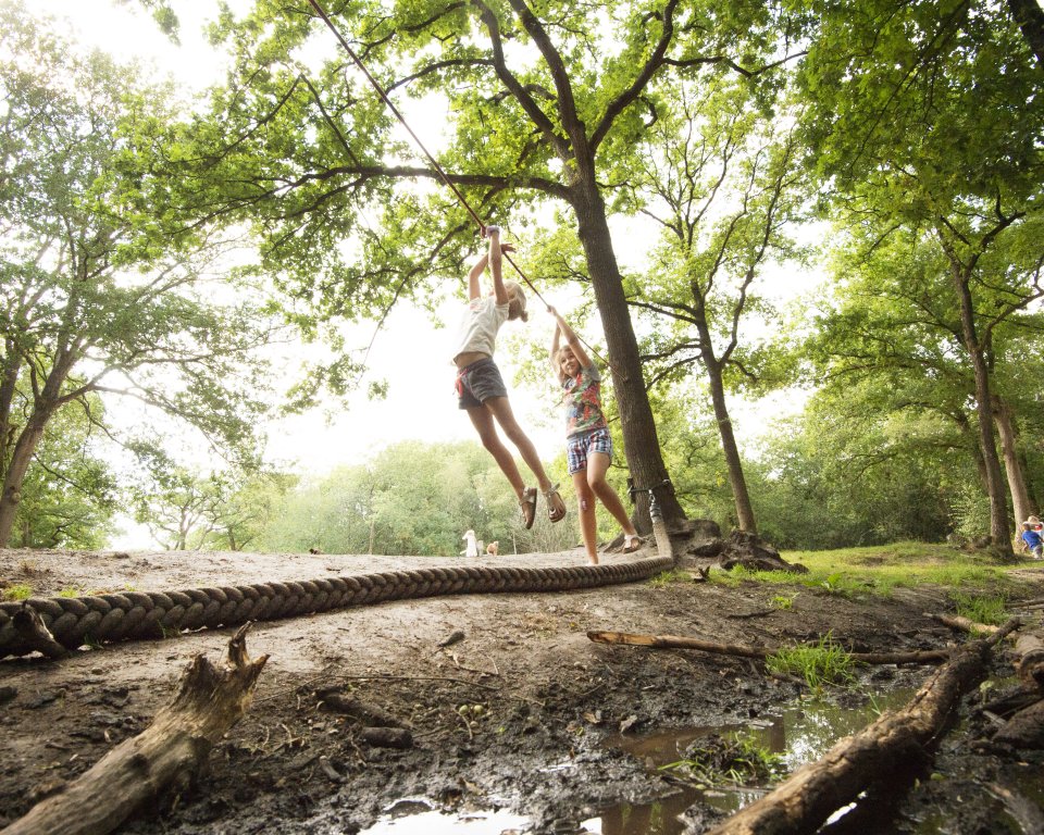 Twee kinderen slingeren aan een touw over een slootje in het bos bij Sparjebird