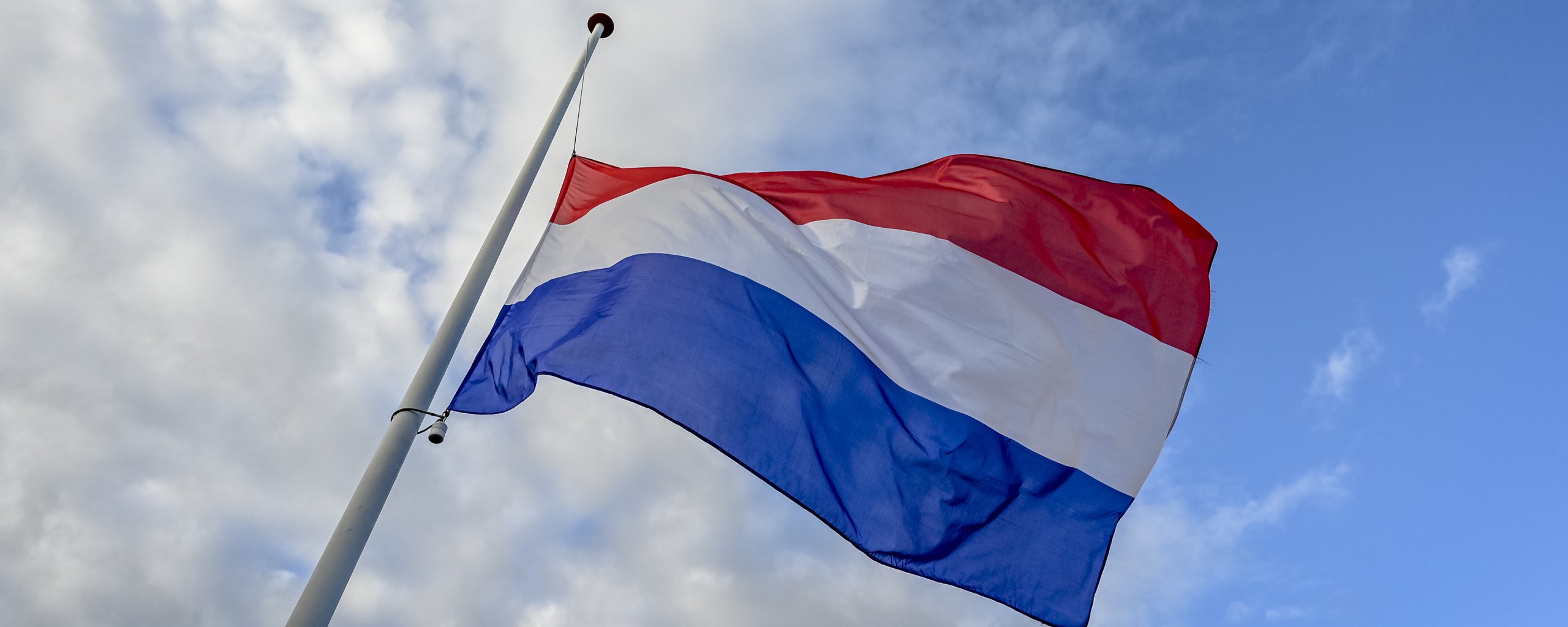 Nederlandse vlag aan een mast, wapperend in de wind tegen een blauwe lucht met wolken.