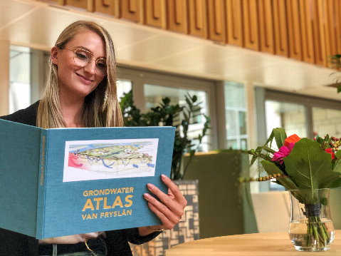 Vrouw met bril en lang blond haar met de blauwe kaft van de atlas in handen met op de achtergrond bloemen