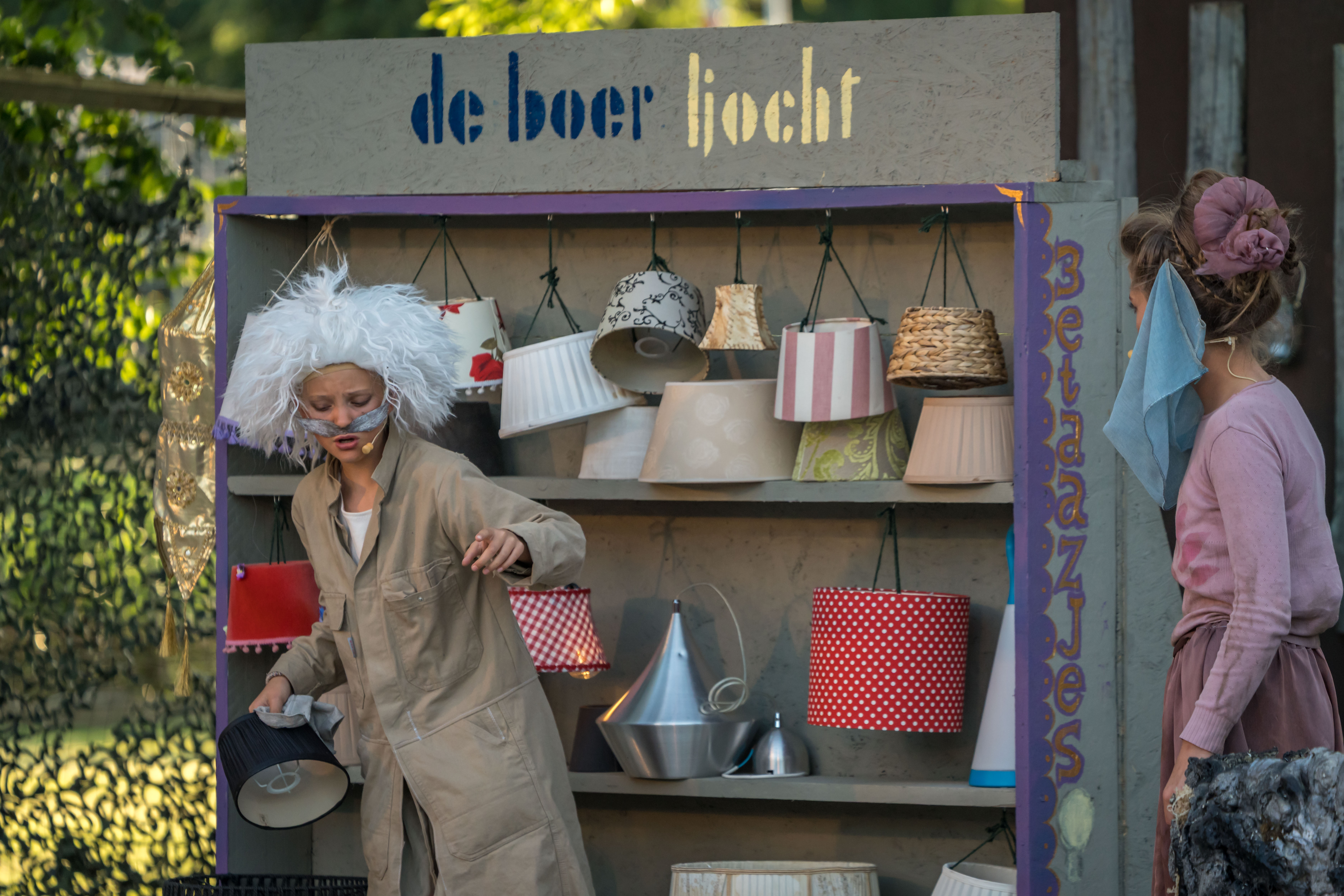 Twee kinderen spelen in de voorstelling Alladin. Een lampenverkoper staat met zijn kar met lampenkappen te spreken.