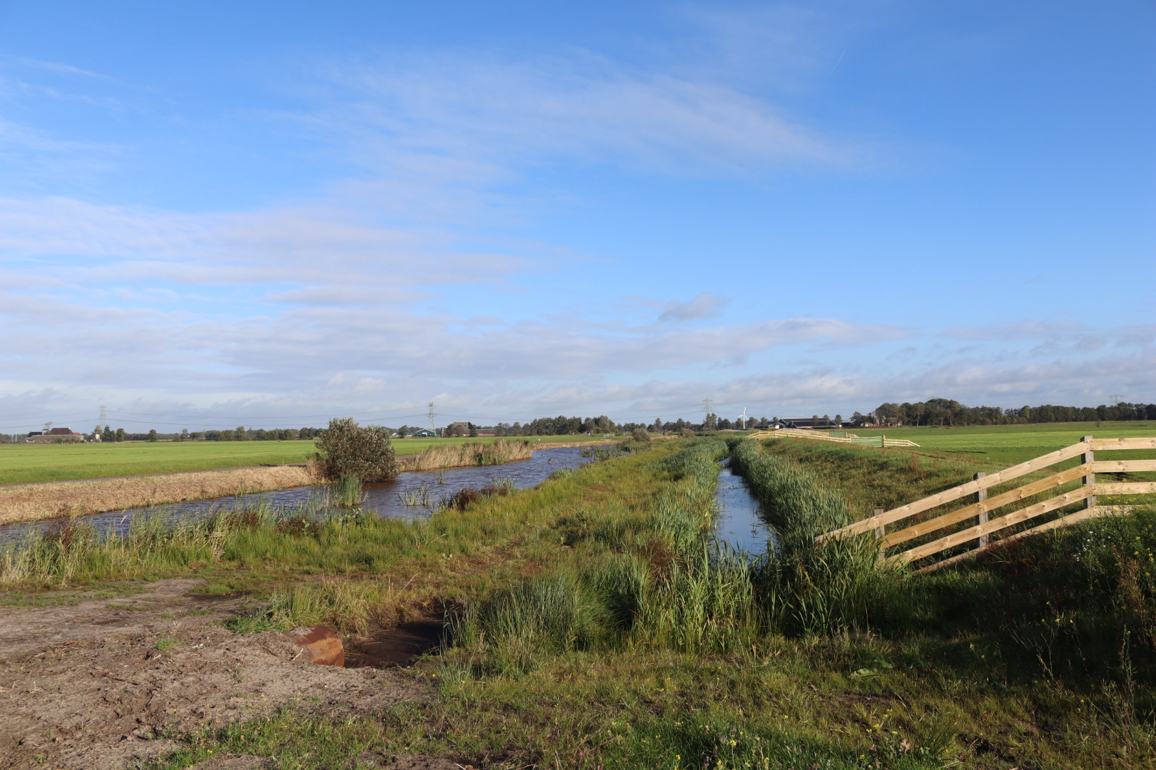 Een weiland waar twee sloten parallel lopen en op de voorgrond een hek.