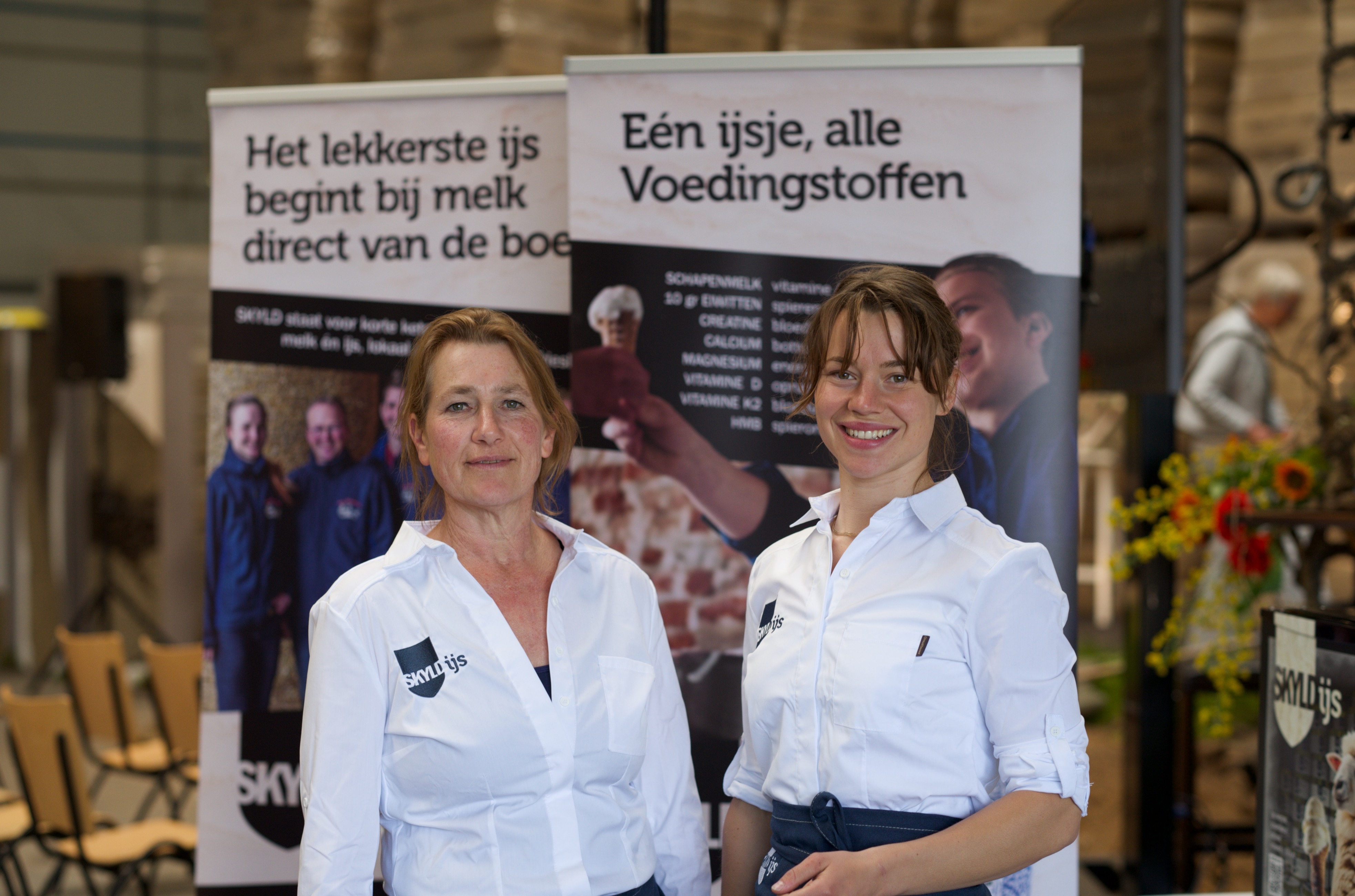 Twee vrouwen met witte bloesjes van SKYLD staan op een beurs.