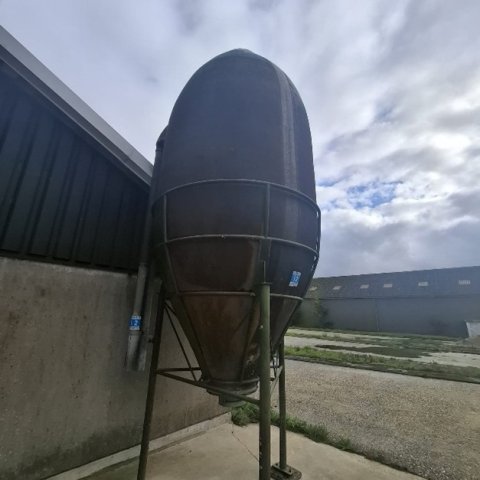 Een silo staat voor een stal op een betonnen vloer met op de achtergrond nog een stal.