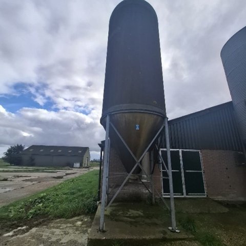Een silo staat voor een stal op een betonnen vloer met op de achtergrond nog een stal.