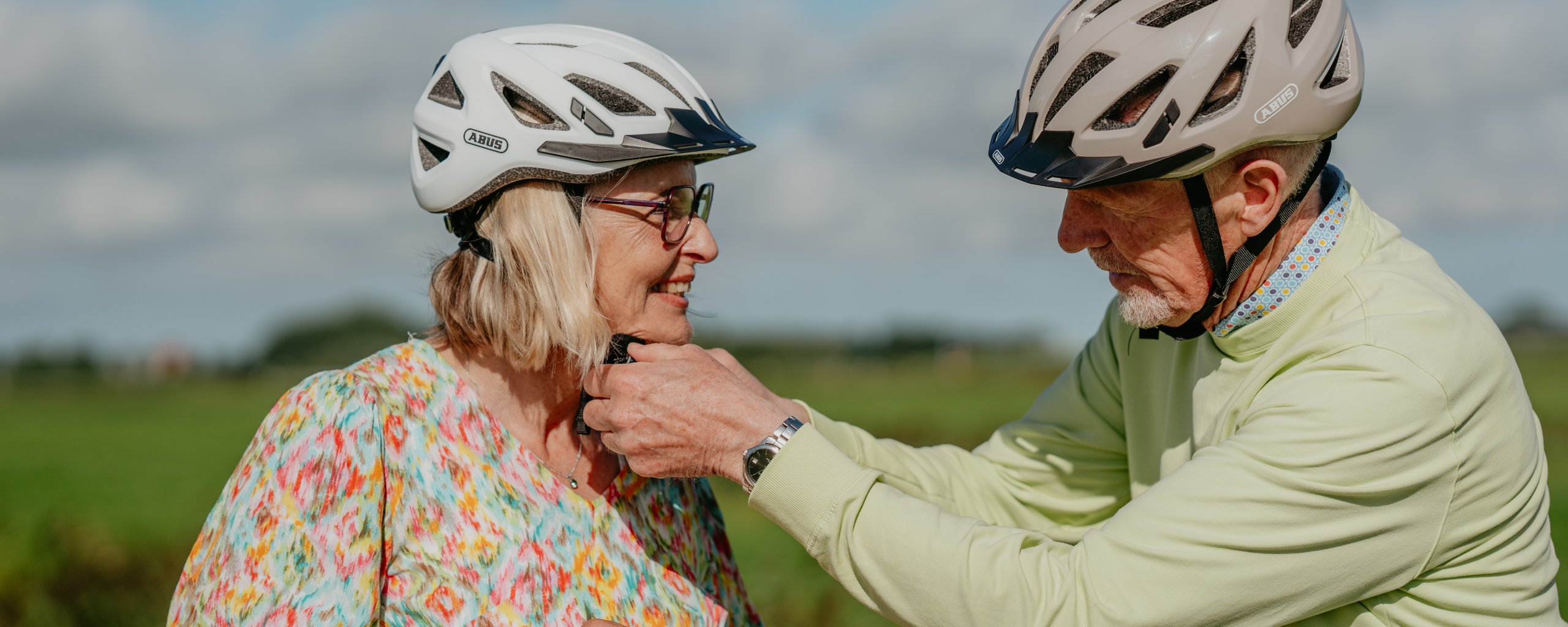 Twee oudere mensen met een fietshelm op. De man doet de helm van de vrouw vast