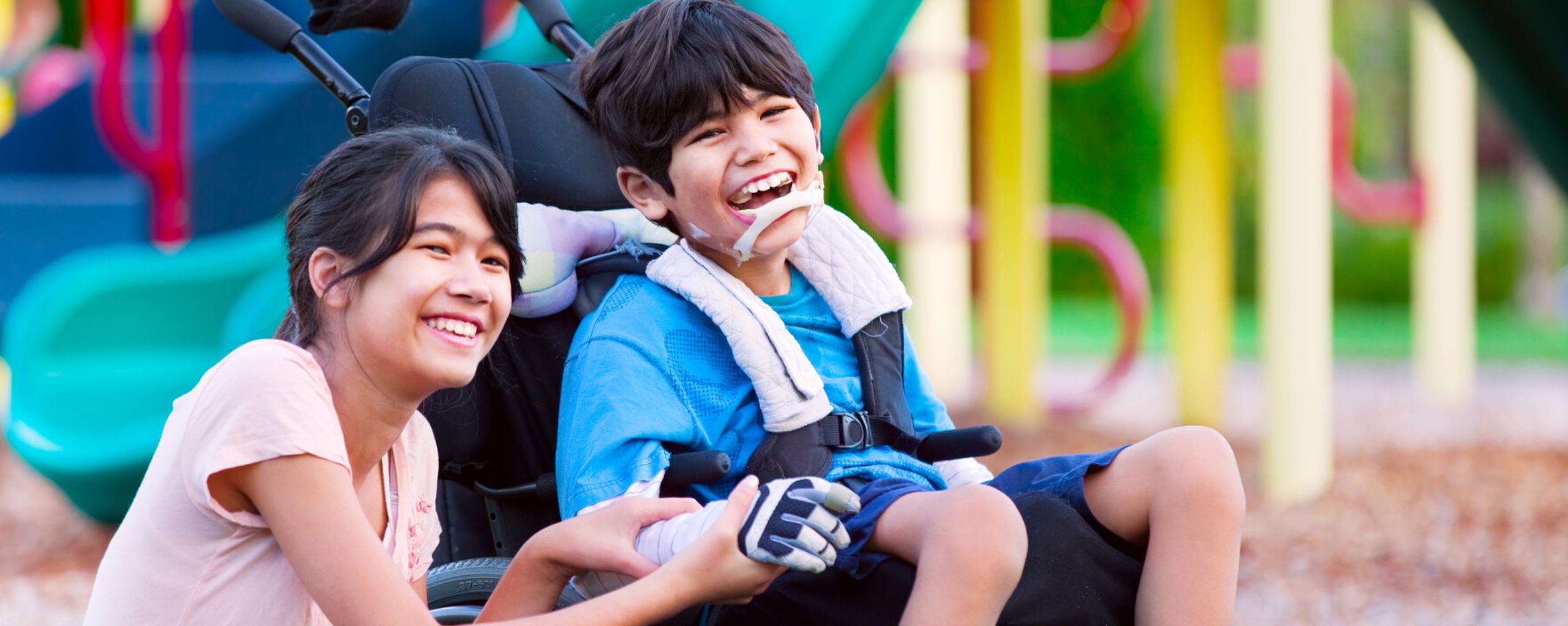 Twee kinderen in een speeltuin; één kind zit in een rolstoel met blauwe kleding en witte handschoenen, terwijl het andere kind naast hem knielt en zijn hand vasthoudt. Op de achtergrond kleurrijk speeltoestel met glijbanen.