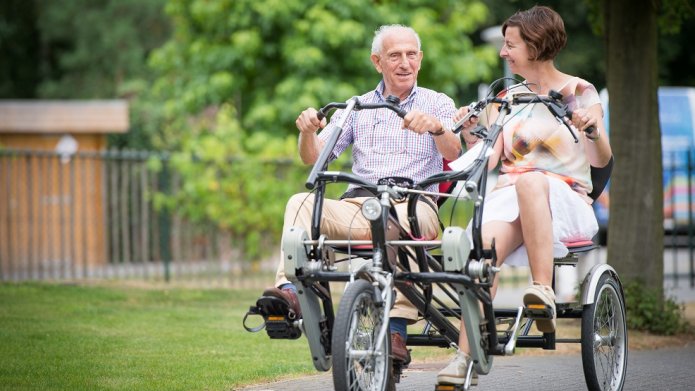 Een oudere heer en een dame rijden samen op een aangepaste tweepersoons fiets.