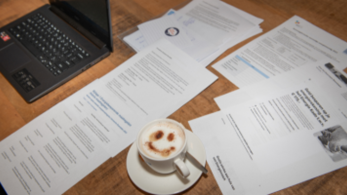 We zien een tafel met formulieren, een laptop en een kopje koffie. 