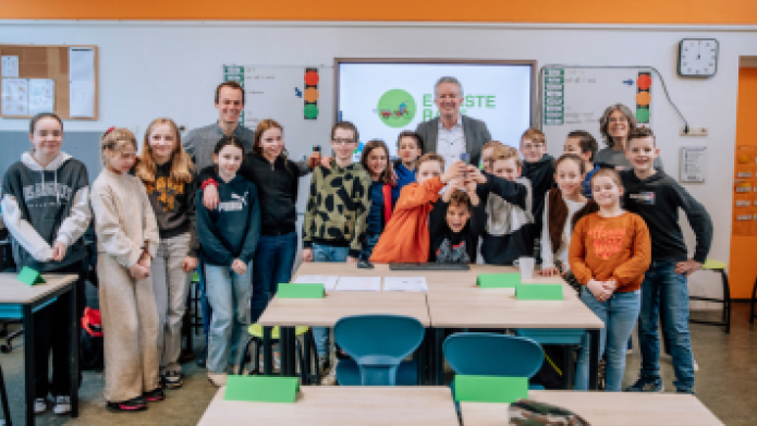 We zien een groep kinderen in een schoollokaal met daarachter een leraar en wethouder Jan Smits. Op de voorgrond tafeltjes en stoelen. 