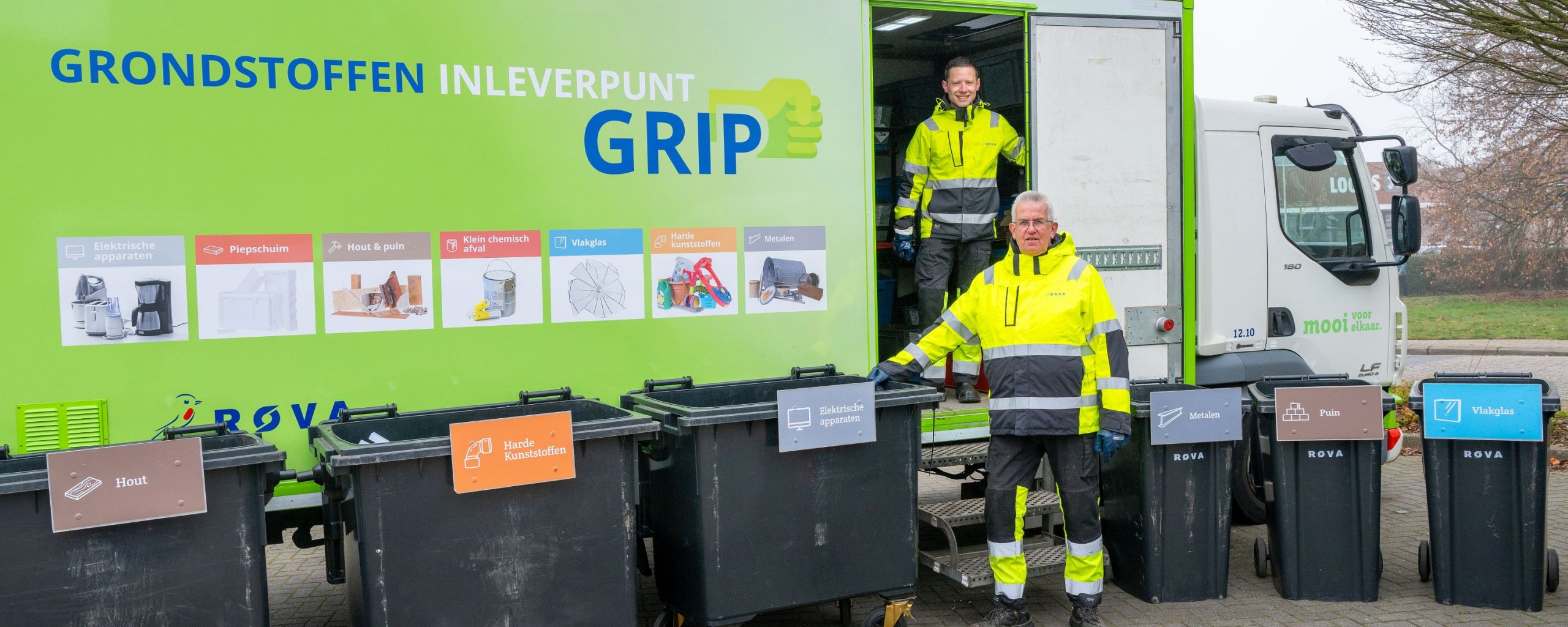 De mobiele inzamelpunt voor grondstoffen is een vrachtwagen. Ervoor staan zeven verschillende afvalcontainers, elk gelabeld met specifieke materialen zoals papier, plastic en metaal. Twee medewerkers in gele veiligheidskleding staan bij de containers en vrachtwagen.