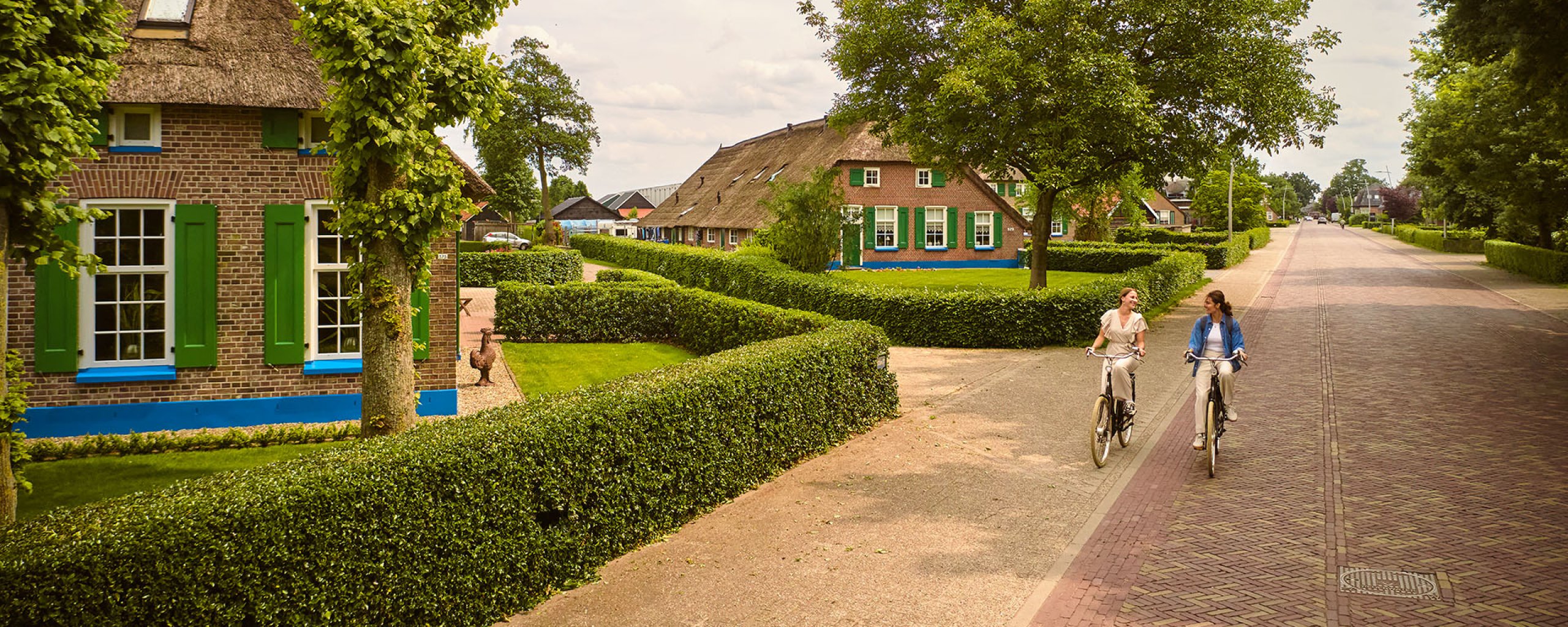 Twee dames fietsen over de Oude Rijksweg langs boerderijen met groene en blauwe kleuren
