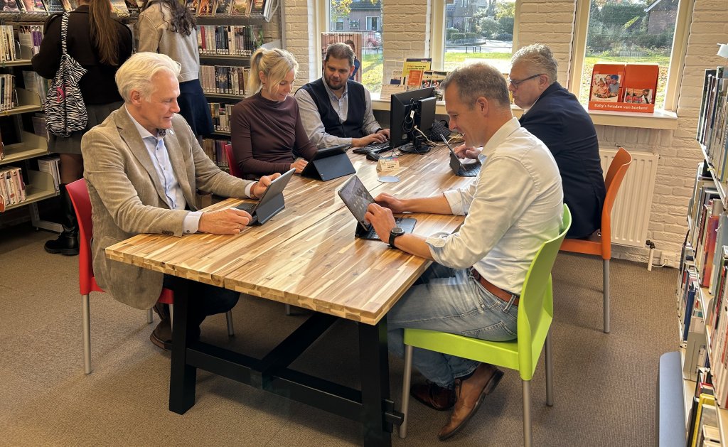 Burgemeester, wethouders en de gemeentesecretaris zitten bij elkaar aan een werkplektafel in de bibliotheek