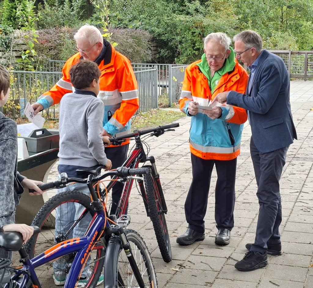 Foto van een fietsactiviteit buiten, waarbij twee medewerkers in fluorescerende oranje werkkleding een jongen begeleiden en papieren controleren. De wethouder kijkt mee over de schouder bij deze controle.