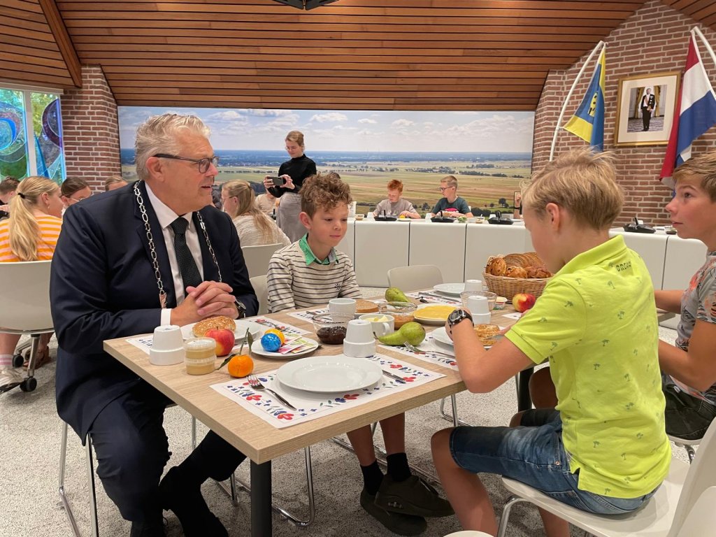 Burgemeester zit aan tafel met kinderen. Op tafel borden, mokken en ontbijtproducten.