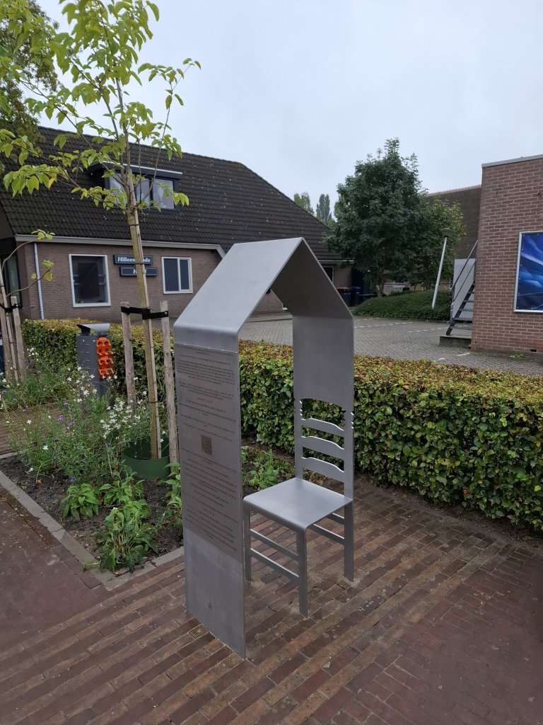 Foto van een metalen informatiezuil voorstellende een stoel dat overgaat in een vorm van een dak van een huis. Gegraveerde tekst vormen het centrale ontwerp, met woningen, struiken en rode bloemen op achtergrond.