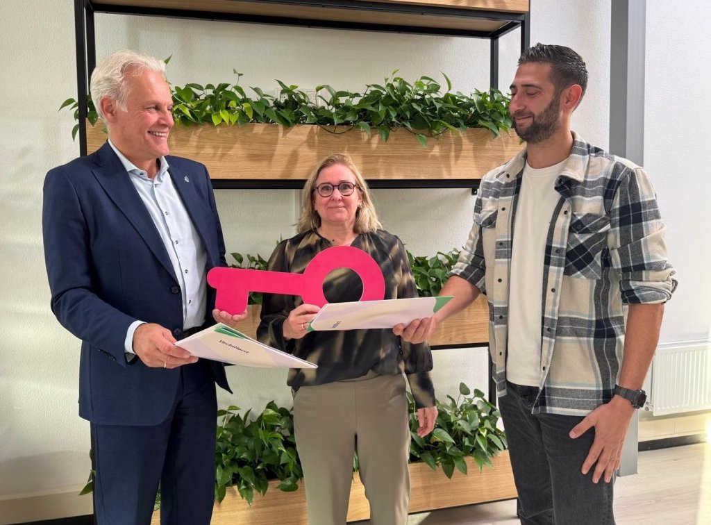 Foto van drie volwassenen tijdens een feestelijke overdracht, waarbij een grote roze sleutel en documenten centraal staan. Links wethouder Mulder, in het midden mevrouw Van Schaik en rechts een medewerker van de woonstichting. Ze poseren lachend voor een houten plantenrekken in een kantoorruimte.
