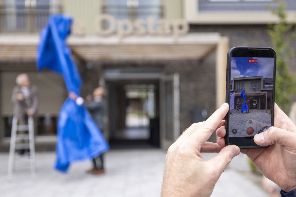 Een blauw doek wordt weggenomen waarmee de naam De Opstap wordt onthuld. In het beeldscherm van een mobiele telefoon is de onthulling goed te zien.