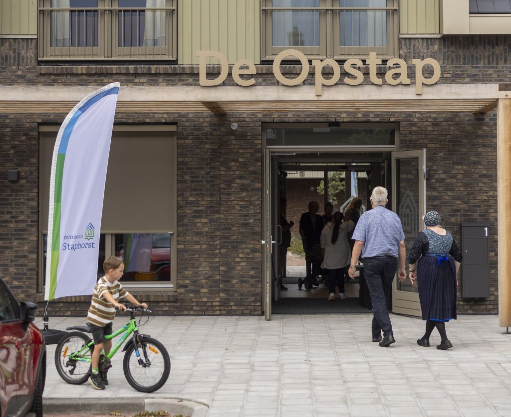 Ingang van het gebouw, echtpaar wandelt naar binnen. Kind op fiets voor beachvlag met logo gemeente Staphorst