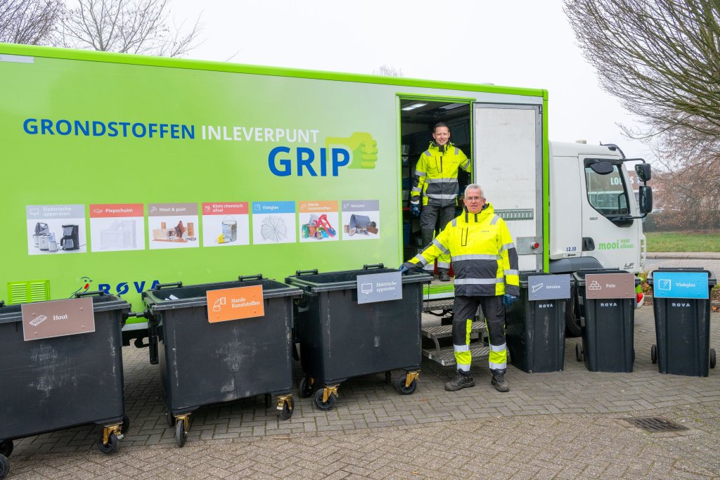 De mobiele inzamelpunt voor grondstoffen is een vrachtwagen. Ervoor staan zeven verschillende afvalcontainers, elk gelabeld met specifieke materialen zoals papier, plastic en metaal. Twee medewerkers in gele veiligheidskleding staan bij de containers en vrachtwagen.