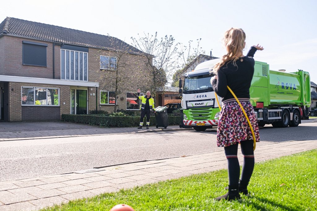 Vuilniswagen van de ROVA in een straat, meisje zwaait naar medewerker van de ROVA die een container weer terugzet