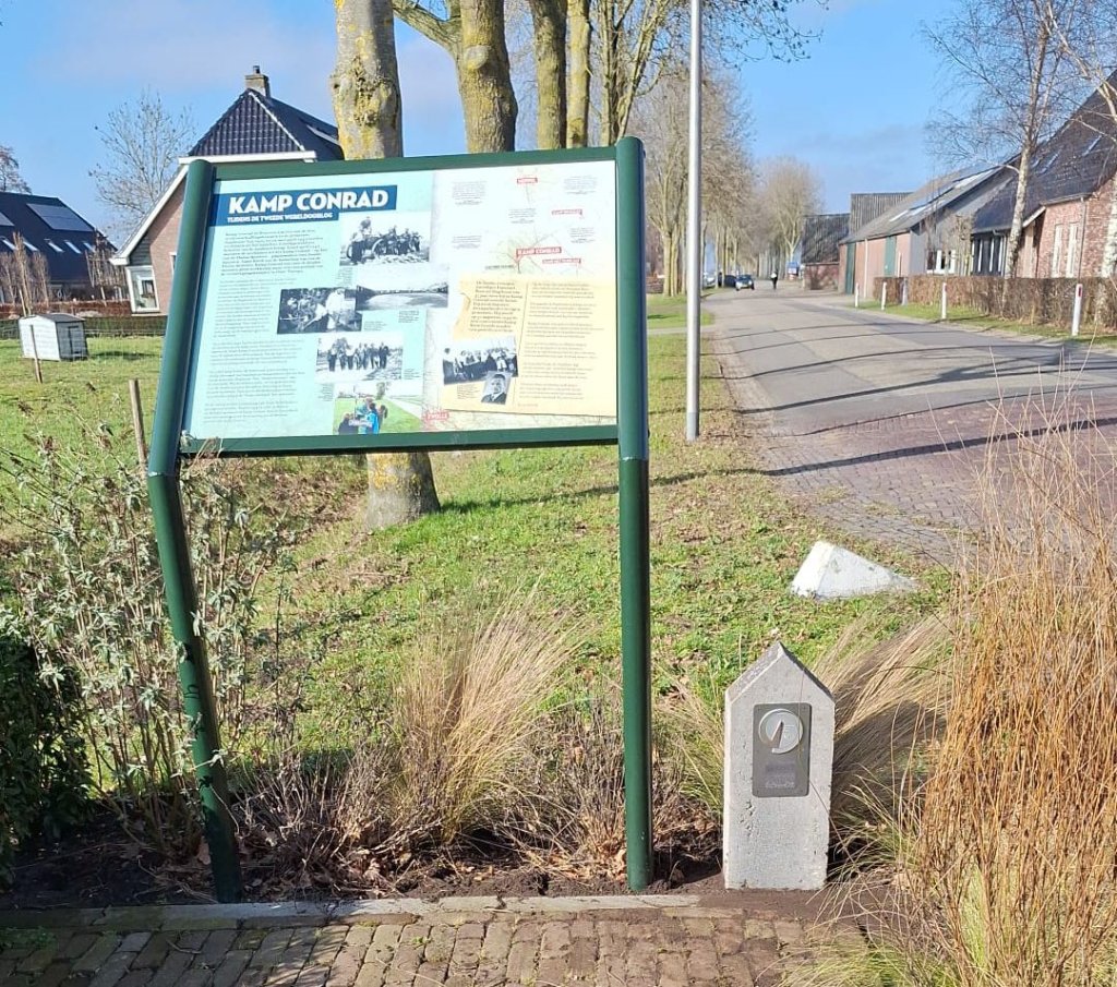 Informatiebord Kamp Conrad, met rechts ervan een grijze markeringspaal met het logo van de Canadian trail