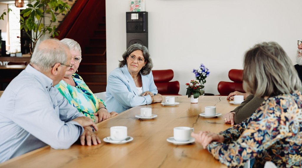 Ouderen rondom een tafel in gesprek. Op de tafel staan kopjes koffie.