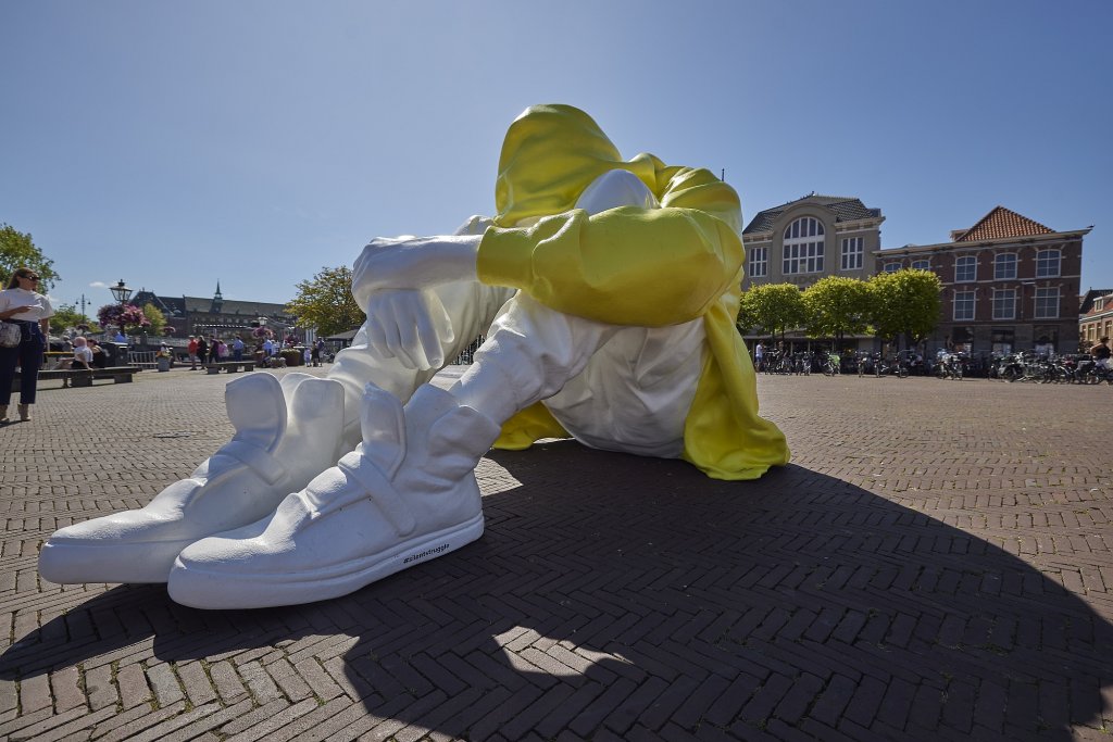 Beel op een plein: voorover gebogen persoon met gele jas en witte broek en schoenen.