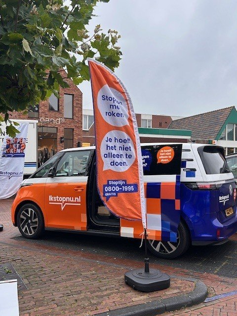 Bus in oranje en blauw met logo ikstopnu.nl. Ervoor staat een beachvlag met tekst Stoppen met roken, je hoeft het niet alleen te doen