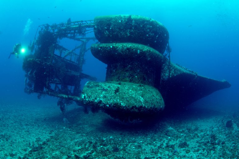 Image shows ship wreckage from behind with diver on the left side showing comparison to the size of the ship vs the size of the diver.