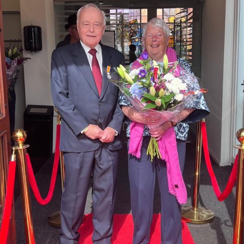 Echtpaar op rode loper voor de raadzaal met bos bloemen en Koninklijk lintje