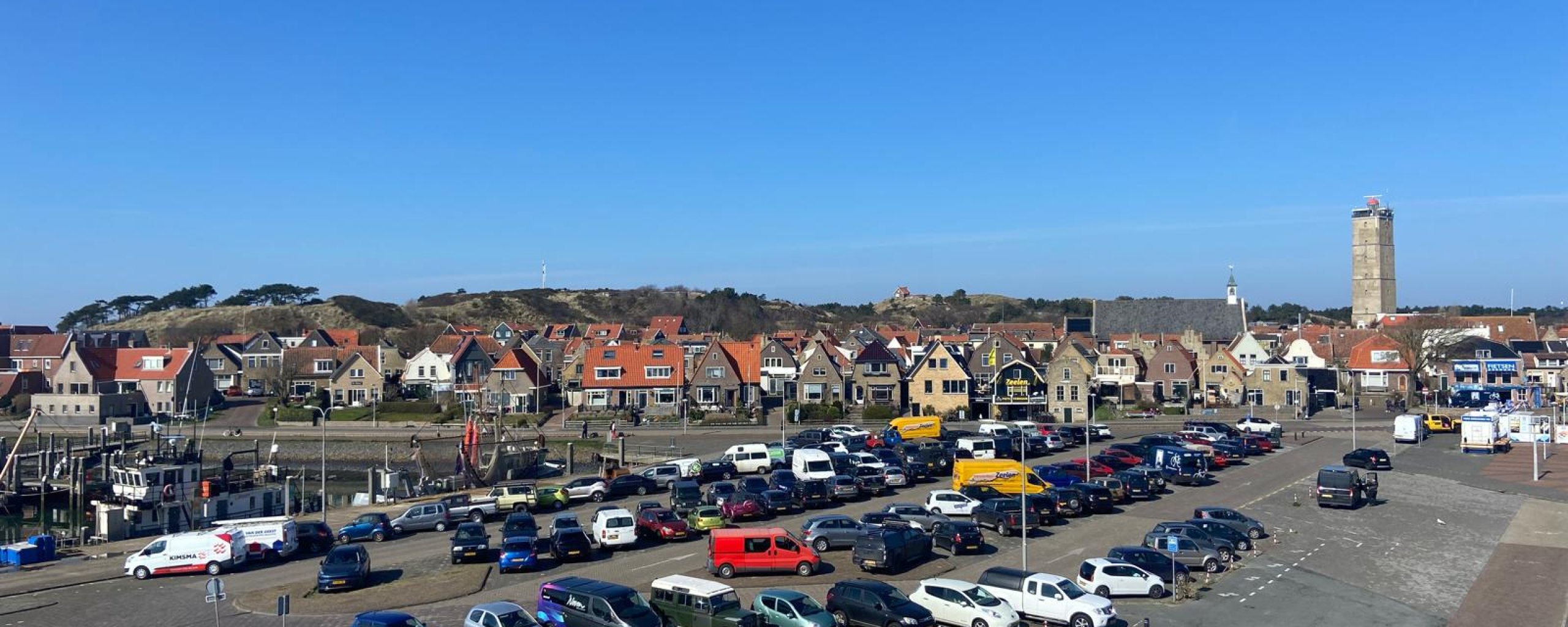 Het Veerhavenplein met allemaal geparkeerde auto's. Daarachter zie je huizen, de duinen en de Brandaris op de achtergrond op een mooie zomerdag..