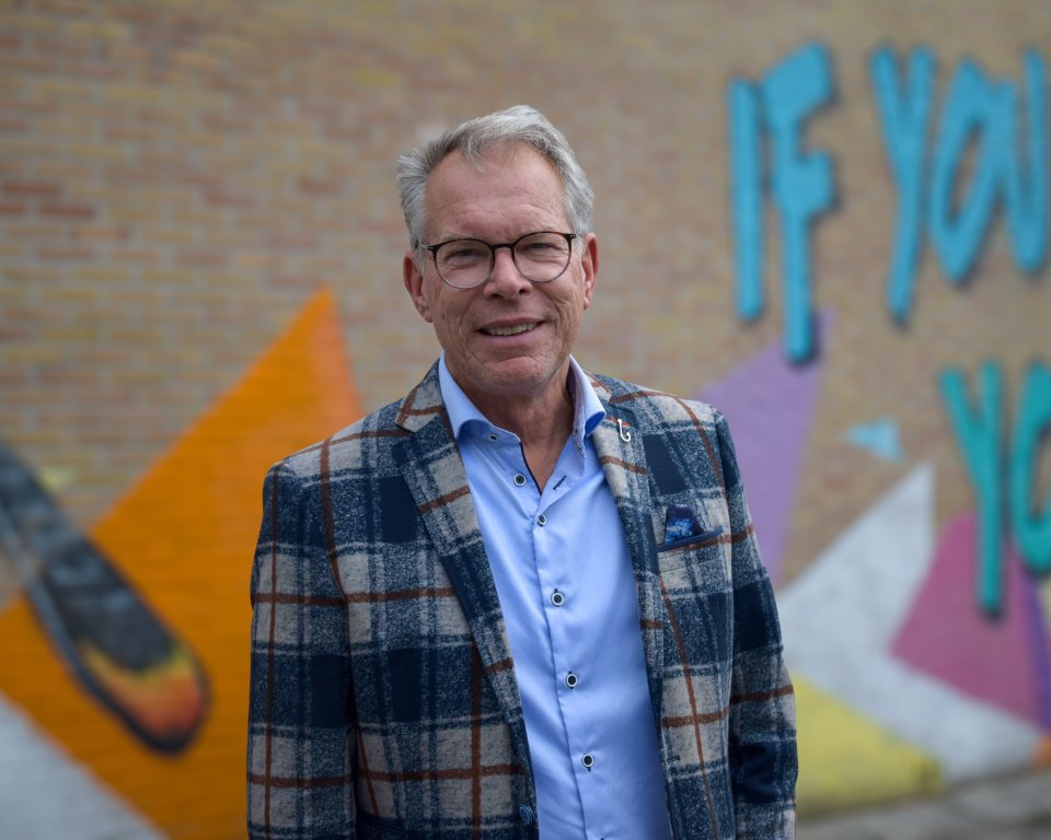 Jan staat buiten voor het schoolgebouw ne kijkt in de camera