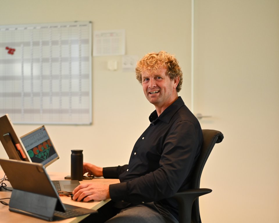 Edzart zit aan tafel achter zijn laptop op de werkvloer en kijkt opzij in de camera. Achter hem hangt een whiteboard met de planning.
