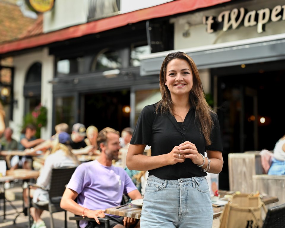 Horecaondernemer Larissa kijkt lachend in de camera terwijl de voor het terras van 't Wapen van Terschelling staat