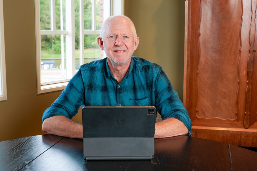Henk zit aan een tafel met zijn laptop en lacht naar de camera